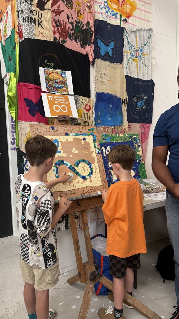 Two children adding mosaic pieces to the SDG 12 panel for Many Hands One Art